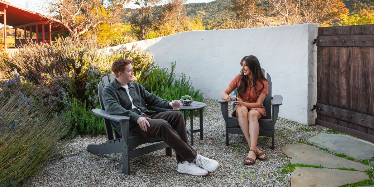 A couple sits in a grey POLYWOOD Classic Adirondack 3 Piece Set in a desert garden surrounded by plants and a white wall.