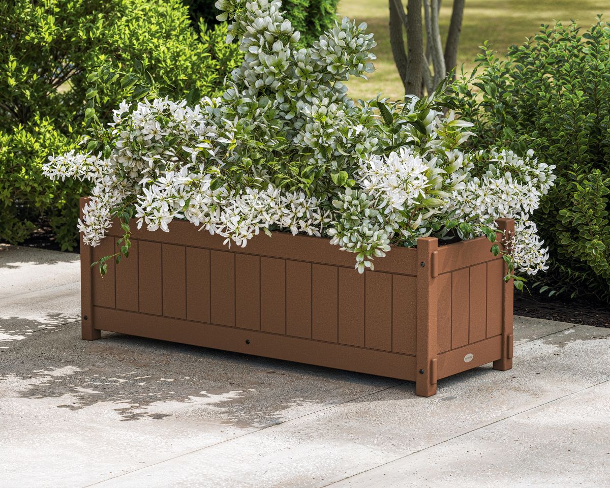 A brown POLYWOOD Vineyard Rectangle Triple Planter Box with white flowers sits on a concrete patio before lush landscaping.