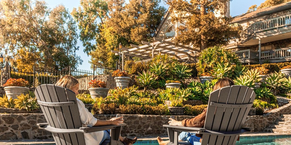 By a pool sit two people in brown POLYWOOD Adirondack chairs facing a landscaped yard with plants, trees, and a pergola. 