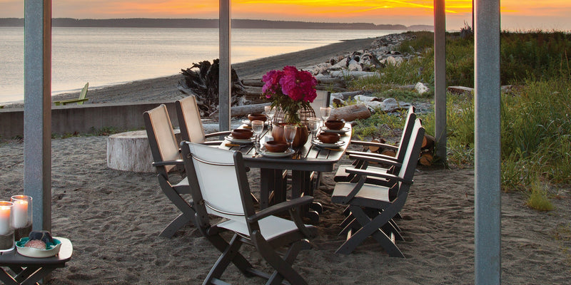 A grey POLYWOOD Coastal Folding Arm Chair 7 Piece Dining Set holds plates, glasses, and flowers on a sandy beach at sunset.