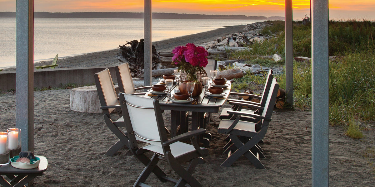 A grey POLYWOOD Coastal Folding Arm Chair 7 Piece Dining Set holds plates, glasses, and flowers on a sandy beach at sunset.