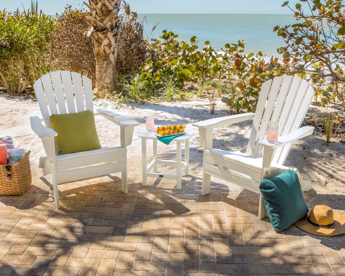 A white POLYWOOD South Beach 3 Piece Folding Adirondack Set sits on a brick patio before a sandy beach overlooking the ocean.