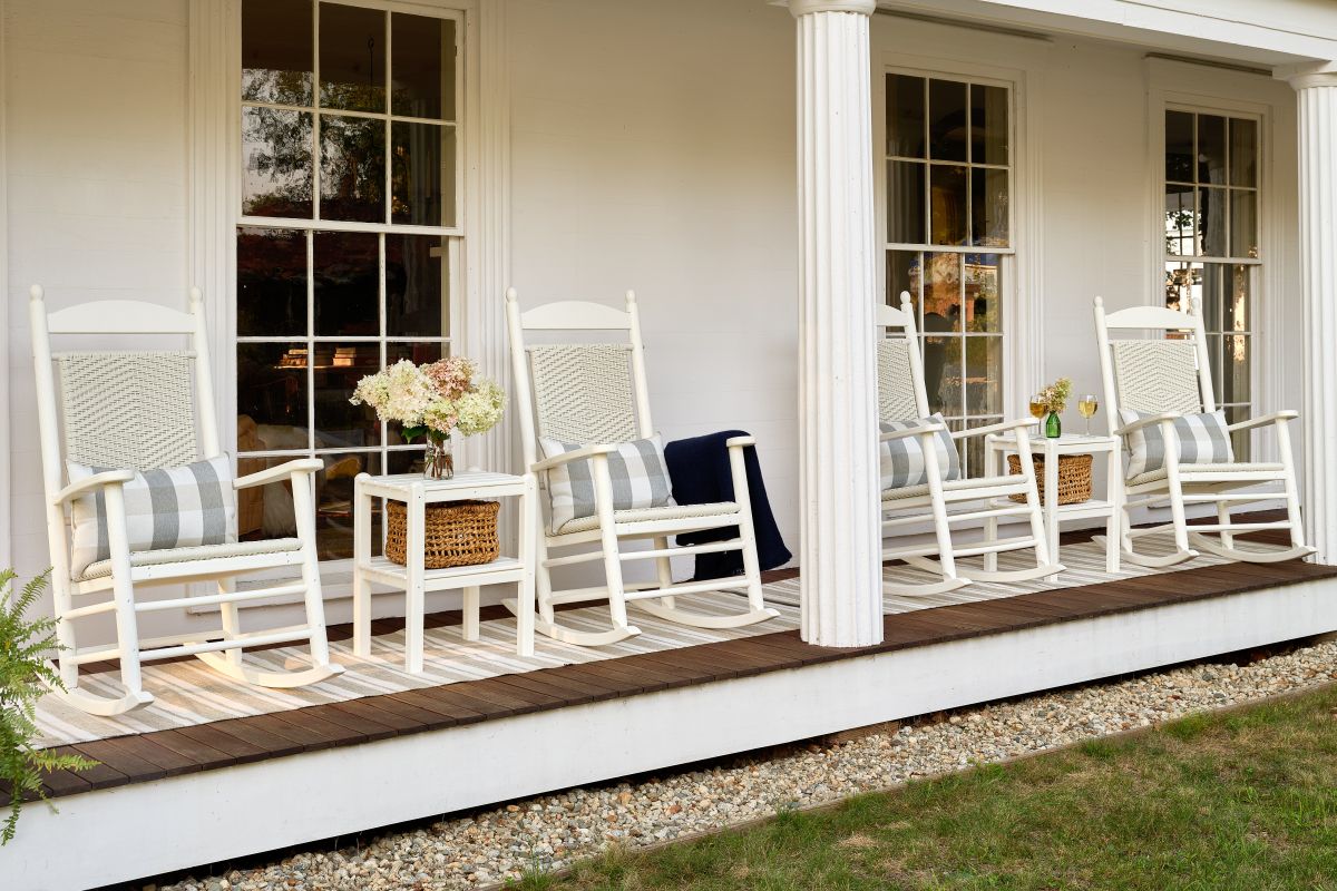 Jefferson Woven Rocking Chairs and Two Shelf Side Tables sit on a wooden covered porch before a white house.