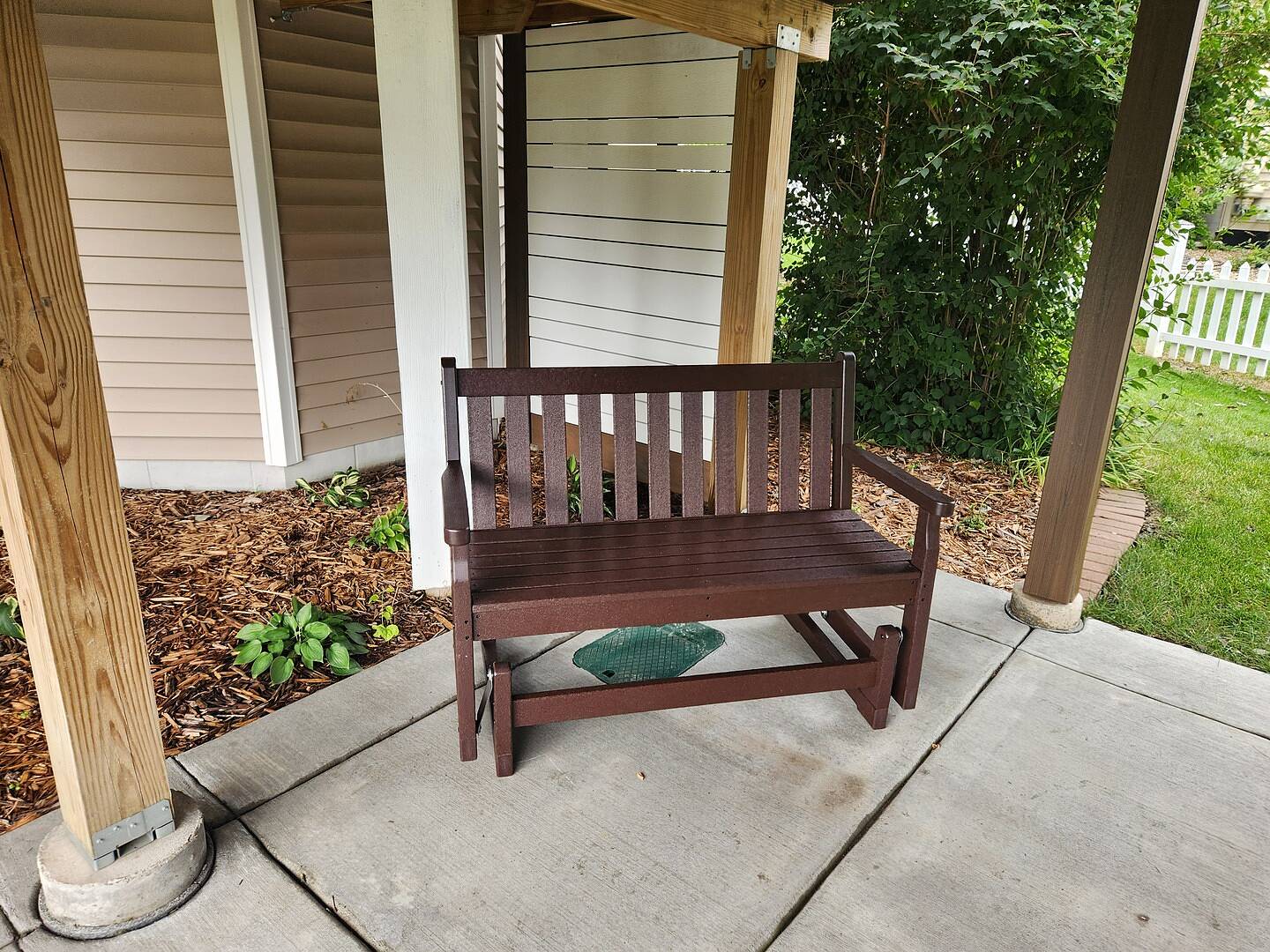 A POLYWOOD Traditional Garden 48 Inch Glider in Mahogany sits on a covered concrete patio before landscaping.