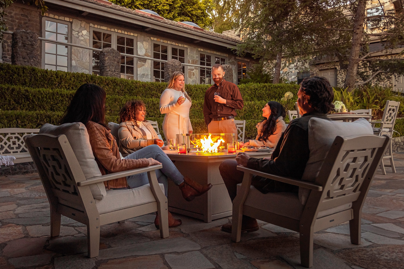 People chat around a POLYWOOD fire pit table in Chinoiserie Deep Seating Chairs on a flagstone patio by a cobblestone house.
