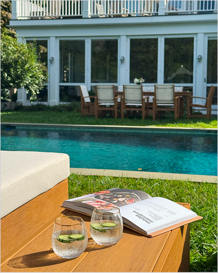 A tan POLYWOOD side table with an open book and water glasses sits by a swimming pool with a dining set on a lawn by a white house with a balcony.