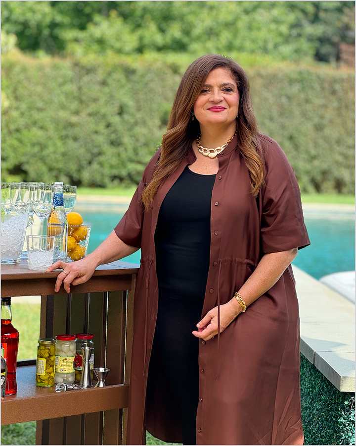 Chef Alex Guarnaschelli stands beside a brown POLYWOOD Vineyard Bar Cart stocked with drinks near a swimming pool.