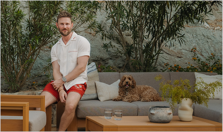 Designer Bobby Berk sits on the arm of a POLYWOOD deep seating sectional with light brown cushions; his dog, Bimini, sits nearby on the sectional. 